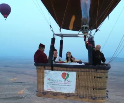 groupe de personnes profitant d’un vol en montgolfière au-dessus d’un paysage désertique. Dans la nacelle, un couple est assis à une table dressée pour un repas, entouré de deux membres d’équipage, dont un en costume traditionnel marocain. Au-dessus d’eux, le brûleur de la montgolfière est en action, projetant des flammes. En arrière-plan, on peut apercevoir une autre montgolfière dans le ciel clair du matin. La nacelle porte une bannière avec l’inscription “Moroccan Sky, ballooning in Morocco - Sky with us”. Cette scène capture un moment de loisir et d’aventure dans le ciel marocain.